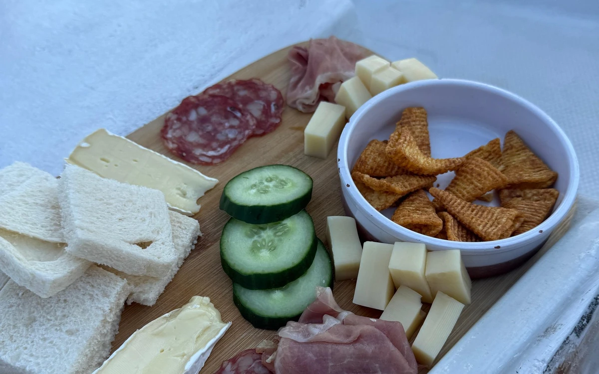 A wooden board with cured ham, salami, cubes of hard cheese, soft cheese, sliced cucumber, white bread and a bowl of crisps