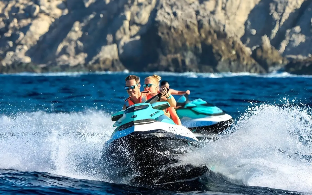 Two riders in sunglasses and life jackets on a jet ski at speed, spray around them, a second jet ski and grey cliffs behind