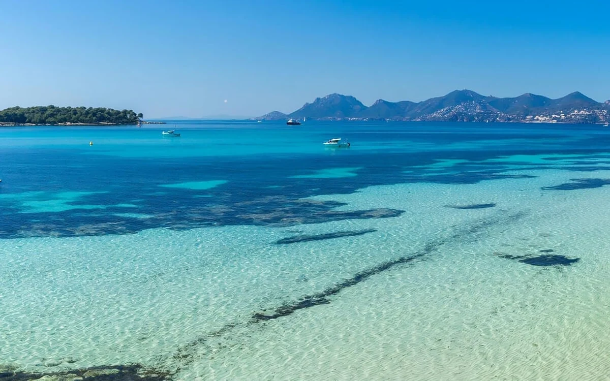 Clear turquoise shallows over sand, small boats moored further out, a wooded islet on the left and hills on the far shore