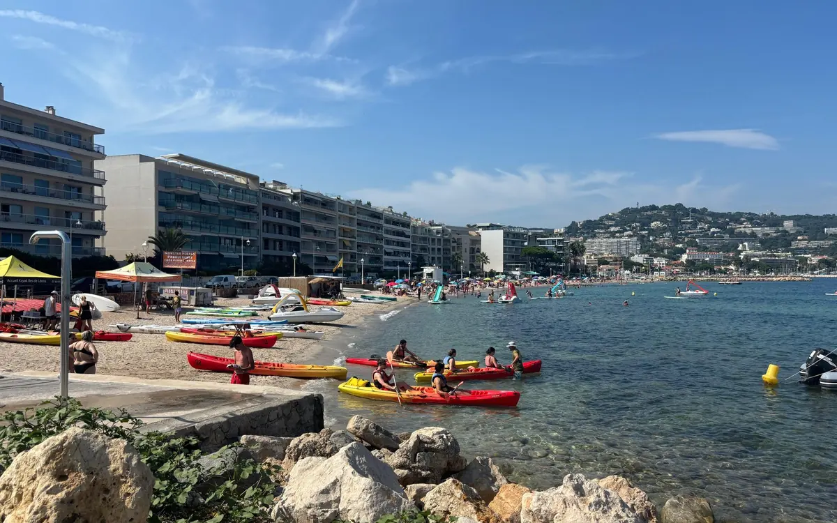 Kayaks at the edge of a sandy town beach, apartment blocks along the shore and hills beyond