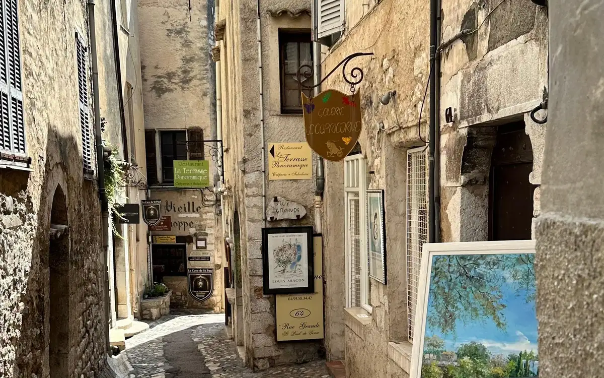 A narrow stone lane in a hilltop village, painted shop signs hanging over the cobbles