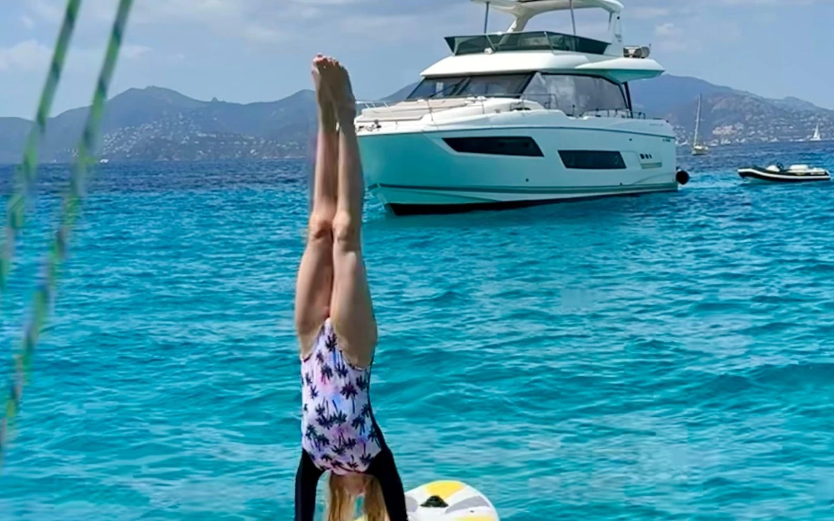 A girl doing a handstand on a paddle board in blue water, a motor yacht and hills behind
