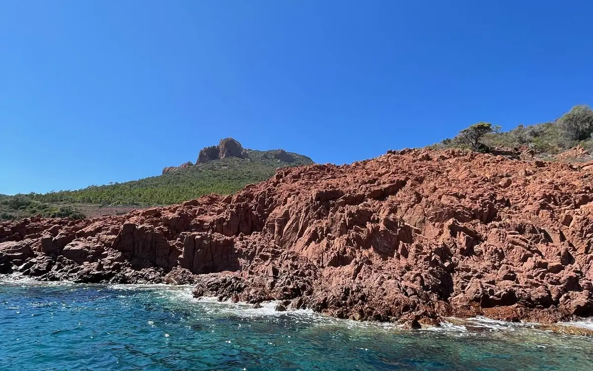 Jagged red rock dropping into blue water, a green hill with a rocky peak behind it
