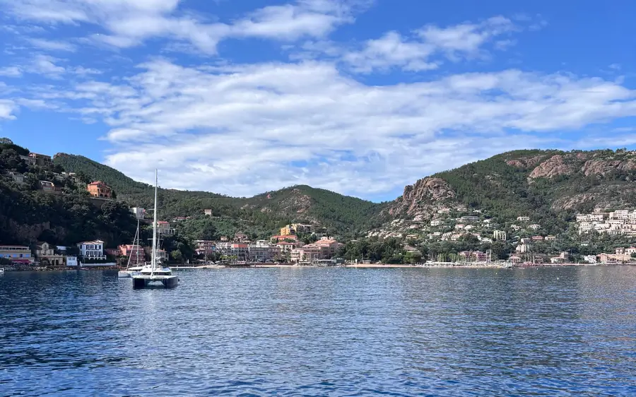 A white catamaran anchored in a blue bay below green hills and a seaside village
