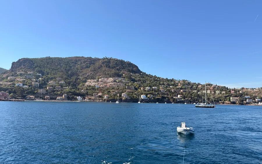 A wooded headland with villas above a blue bay, a small tender and a sailboat offshore