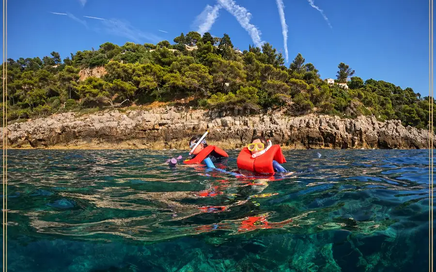 Two snorkellers in red buoyancy vests in clear green water below a rocky shore