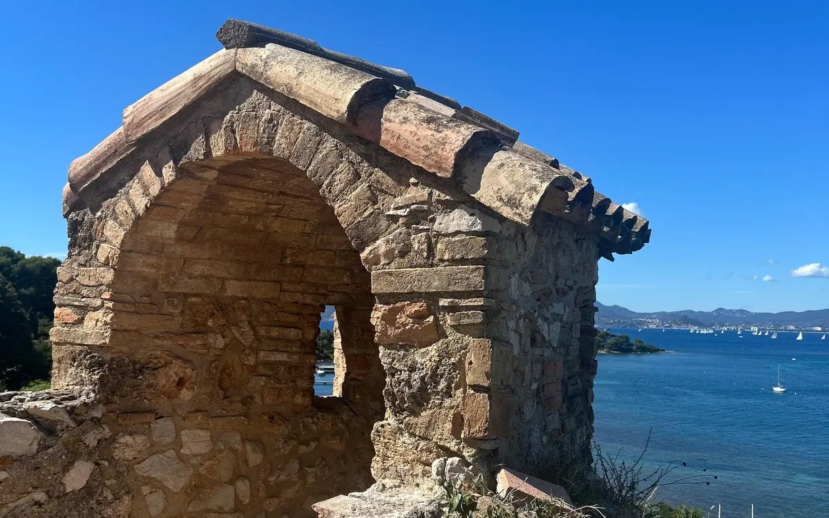 An old stone arch with a tiled roof above the sea, sailboats and the coast beyond