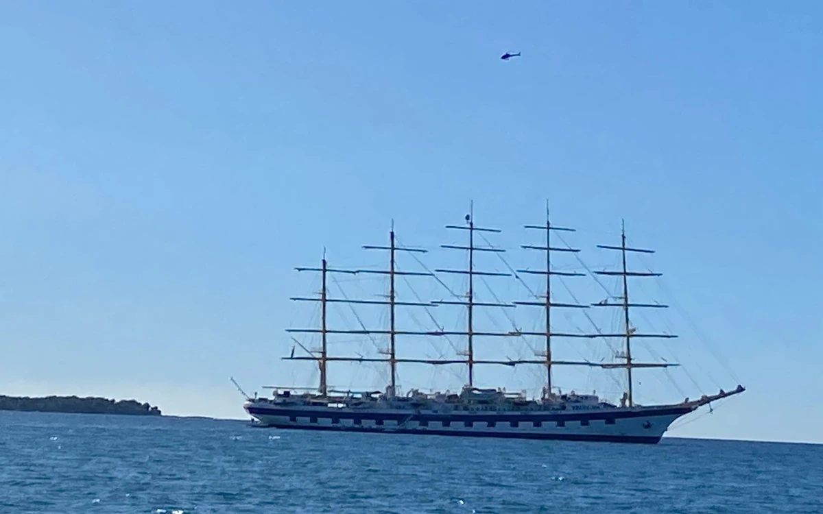 A five-masted sailing ship lying off a low wooded shore, a helicopter in the sky above