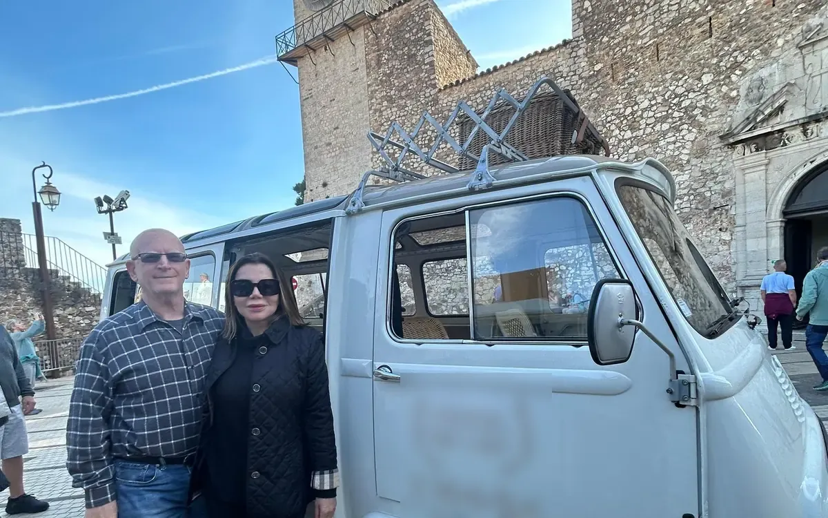 Two visitors standing beside a white vintage minibus with a roof rack, a stone church tower behind